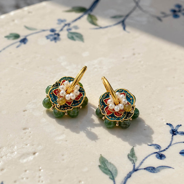 Moss Garden convertible hoop earrings with hand-painted enamel, pearls and green gemstones displayed on a cream-colored ceramic surface with blue floral painted motifs.