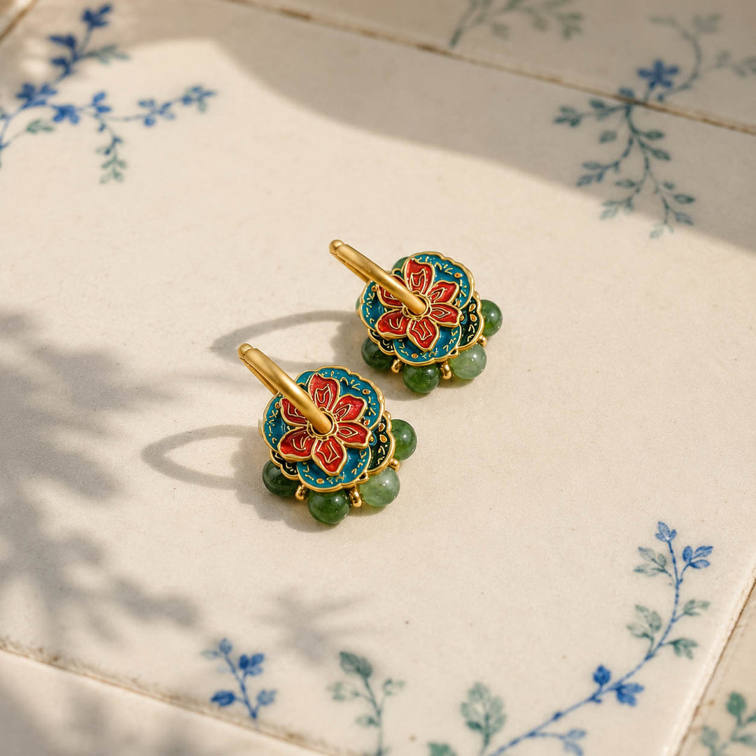 Convertible hoop earrings with red teal enamel flowers and moss-green Canadian Jade beads, resting on a white ceramic tile with blue floral patterns.