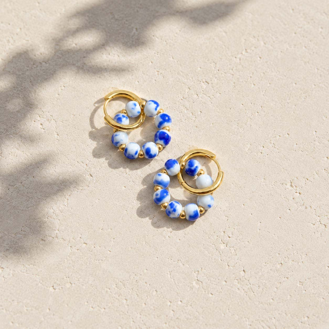 A pair of gold hoop earrings with blue and white porcelain beads, photographed on a light beige textured surface with soft leaf shadows.