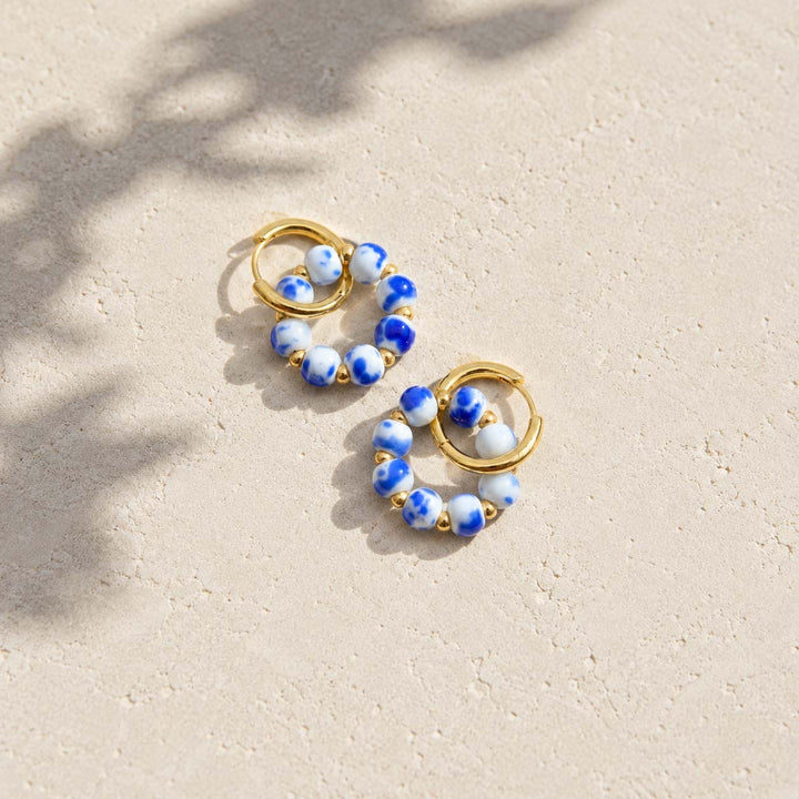 A pair of gold hoop earrings with blue and white porcelain beads, photographed on a light beige textured surface with soft leaf shadows.