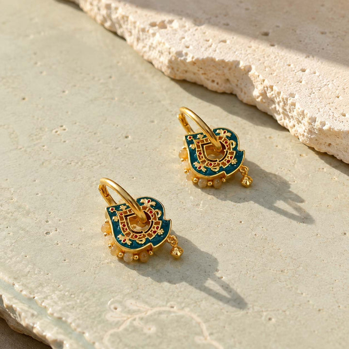 Teal hand-painted enamel convertible hoop earrings with Yellow Jade bead accents, resting on a textured beige stone surface.