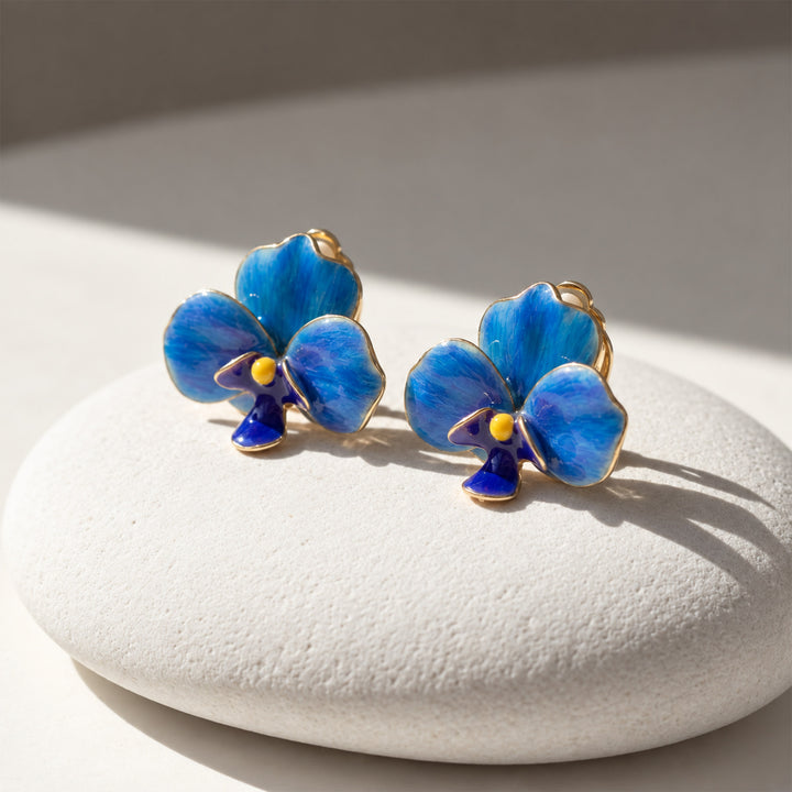 Blue orchid clip-on earrings with hand-painted enamel petals displayed on a sunlit cream stone surface.
