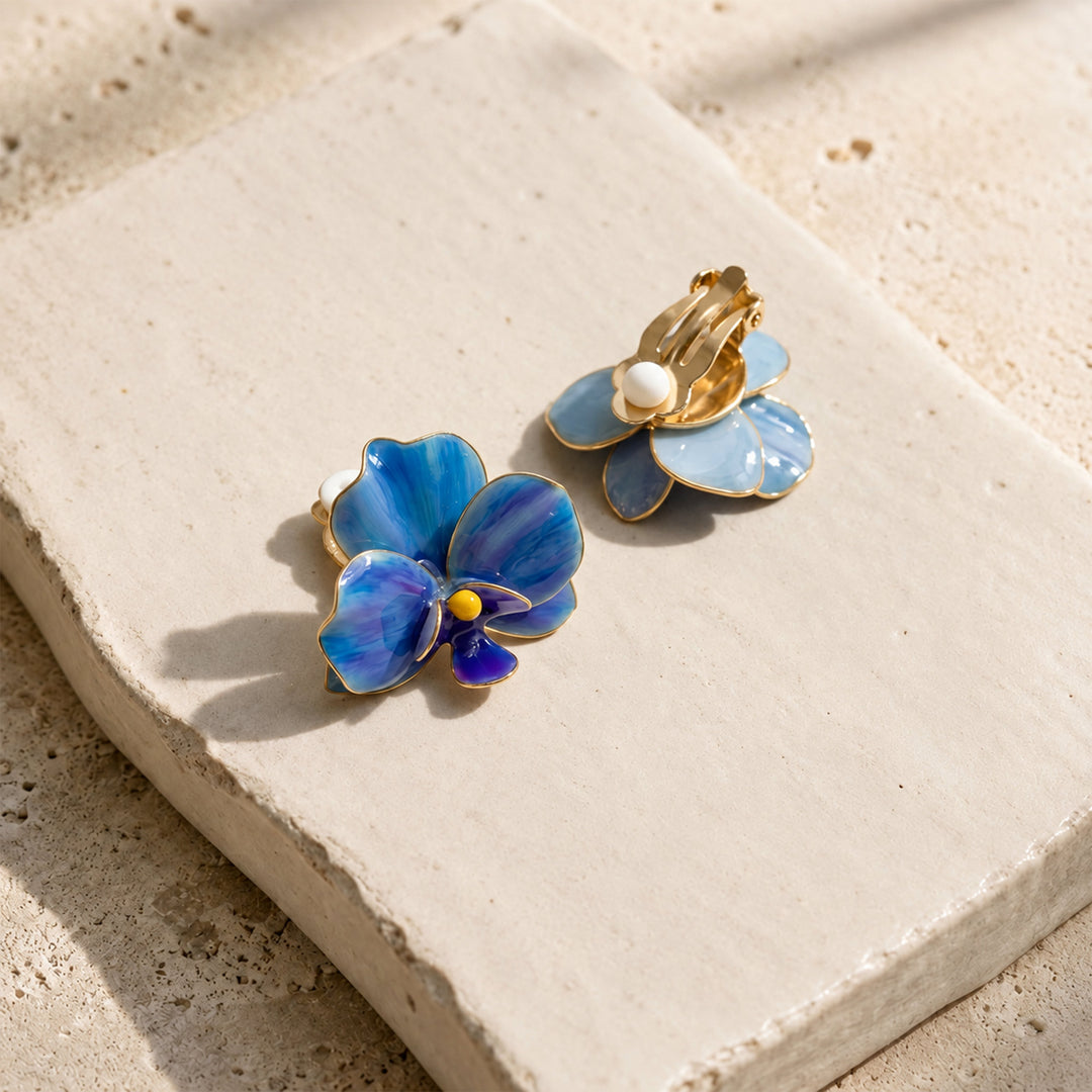 A pair of gold-tone blue orchid earrings with blue and light blue enamel petals, displayed on a textured beige stone tile.