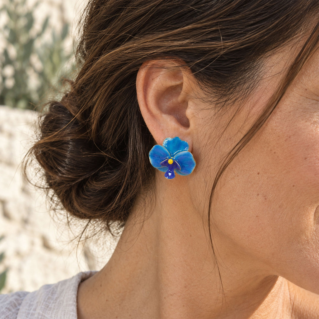 Model wearing hand-painted blue orchid clip-on earrings outdoors against a pale stone background.