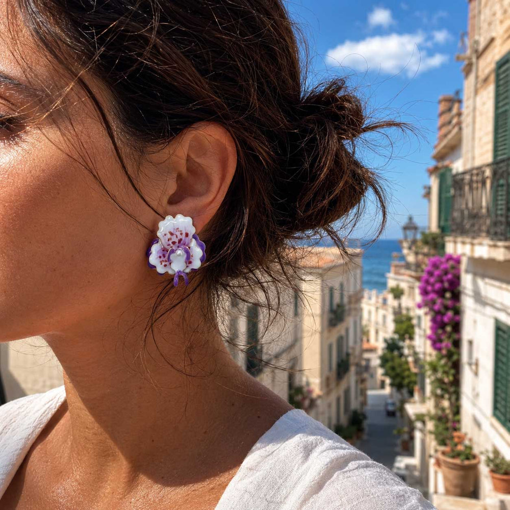 Woman wearing purple orchid clip-on earrings featuring hand-painted enamel petals and 18K gold-plated floral details on a Mediterranean street.