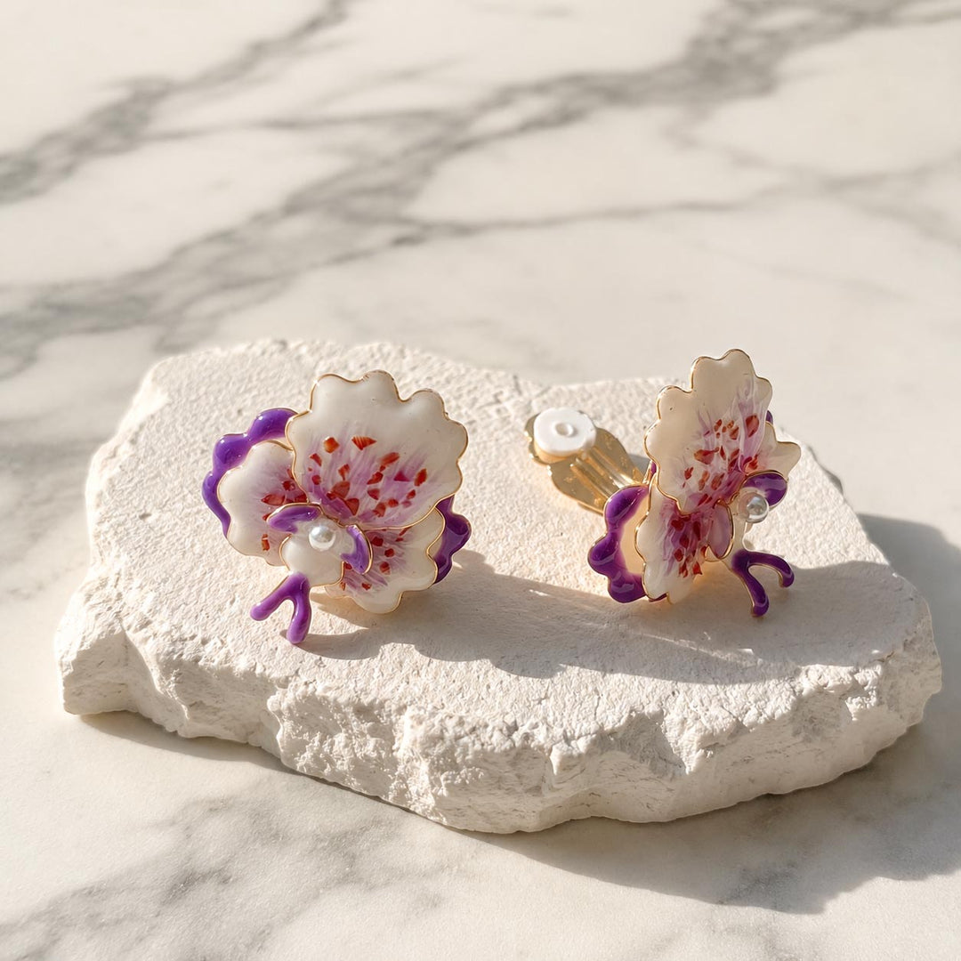 Purple orchid clip-on earrings with hand-painted enamel petals displayed on a textured white stone surface.