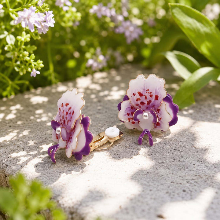 A pair of gold-toned stud earrings shaped like white orchids with purple and red accents, resting on a light stone surface outdoors with green leaves and small purple flowers in the background.