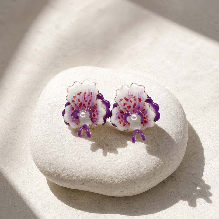 A pair of handcrafted orchid stud earrings with white petals, purple accents, and red speckles, resting on a smooth white stone under warm sunlight.
