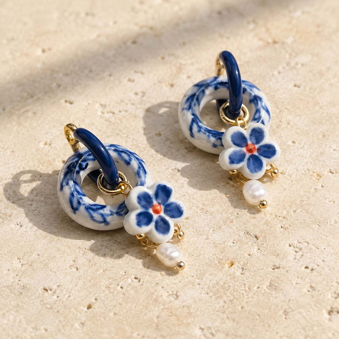 A2K handcrafted convertible hoop earrings featuring handpainted porcelain and freshwater pearls with 18k gold-plated hardware, photographed on a light beige textured surface.