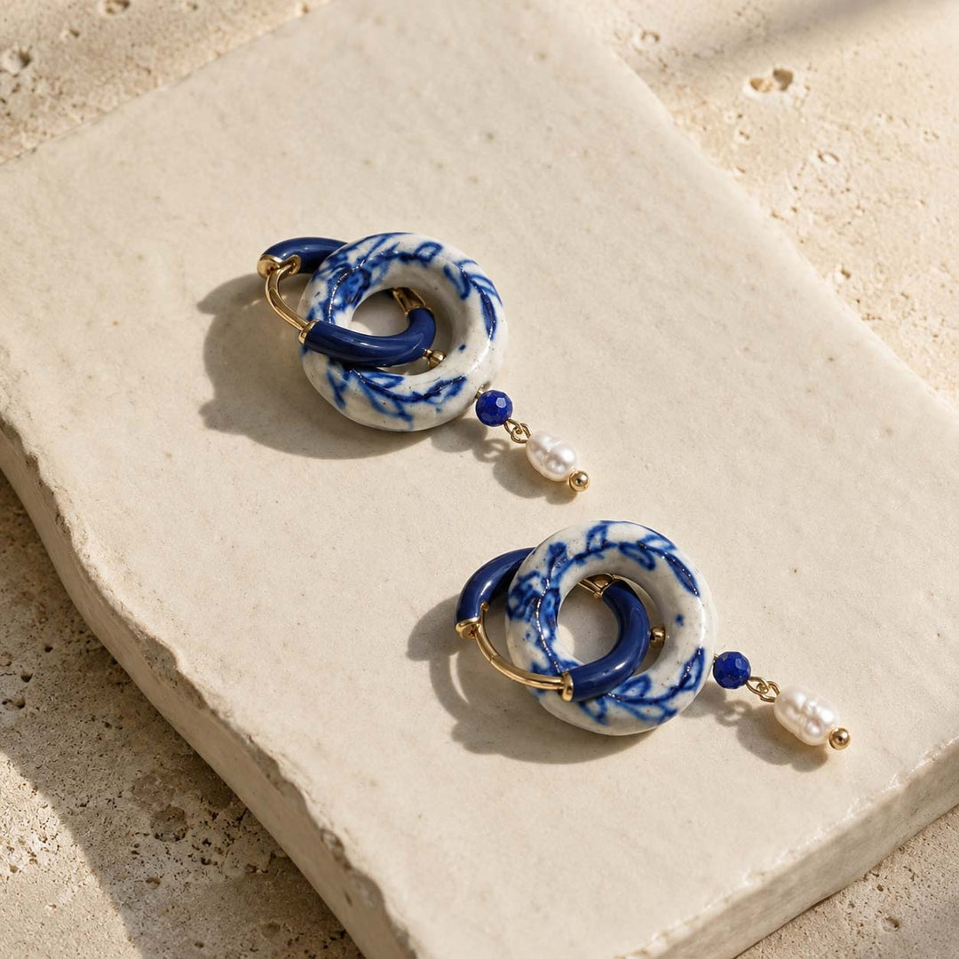 A2K convertible hoop earrings featuring blue and white porcelain with a navy enamel drop, lapis bead, and freshwater pearl, displayed on a textured beige stone tile.