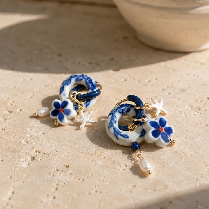 A pair of handmade porcelain hoop earrings with blue floral details, gold-tone hardware, and dangling fresh water pearl, displayed on a textured beige stone surface.