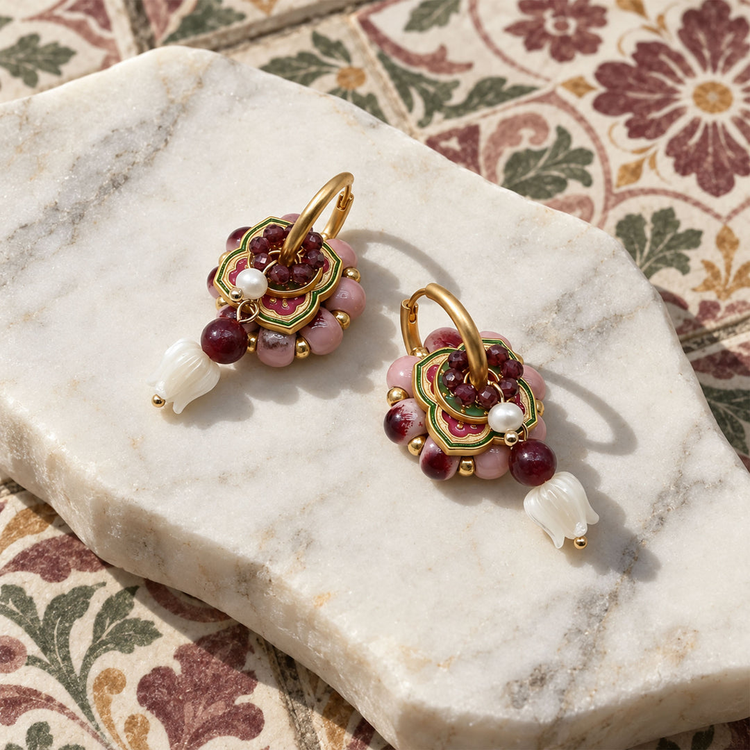 Garnet and pink porcelain convertible hoop earrings with hand-painted enamel floral charms, displayed on a white marble slab over a floral patterned tiles.