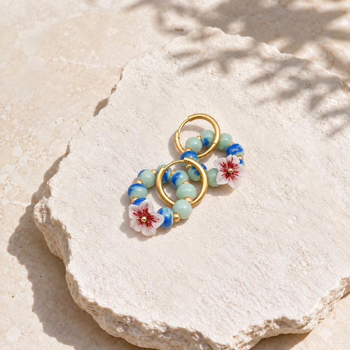Convertible hoop earrings with hand-painted blue-green porcelain beads and sakura flower charms on pale textured stone.