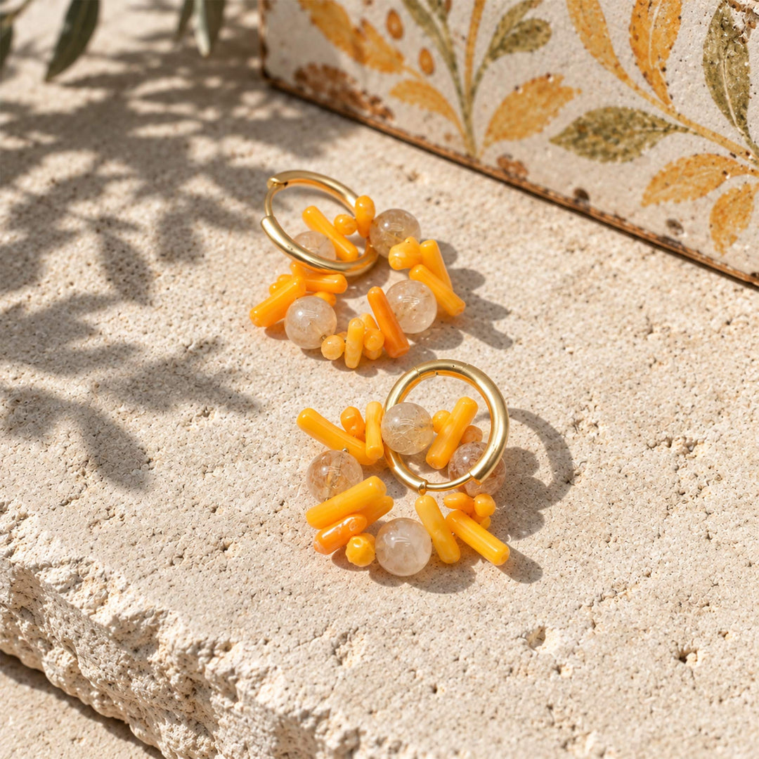 A pair of gold hoop earrings with Citrine and coral displayed on a textured beige stone surface with leaf shadows.
