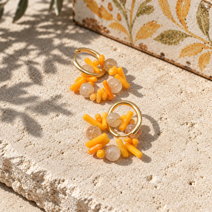 A pair of gold hoop earrings with Citrine and coral displayed on a textured beige stone surface with leaf shadows.