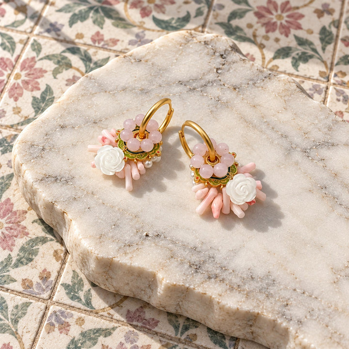 A pair of gold hoop earrings with pink and white floral charms, displayed on a polished stone slab over a floral tile surface.