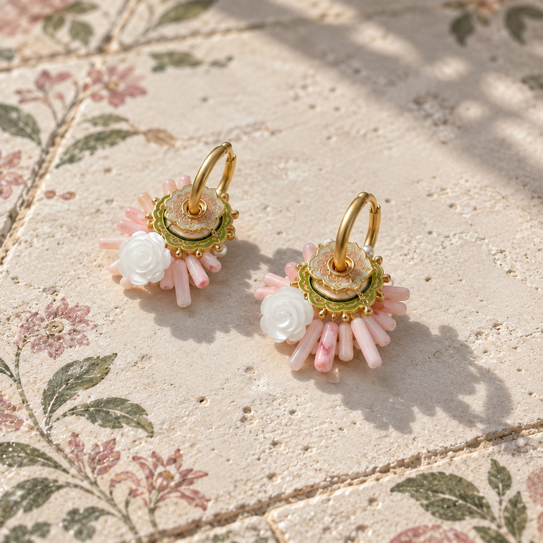 Pink quartz and coral convertible hoop earrings with hand-carved mother-of-pearl roses, resting on a textured beige stone surface with a floral patterned background.