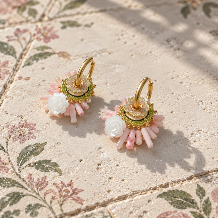 Pink quartz and coral convertible hoop earrings with hand-carved mother-of-pearl roses, resting on a textured beige stone surface with a floral patterned background.