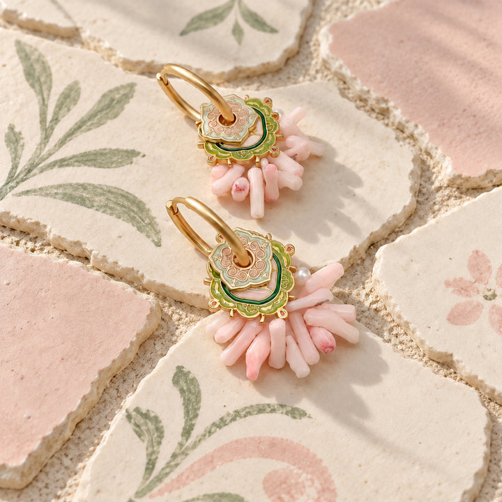 A pair of gold hoop earrings with layered floral charms in green, pink, and gold, hanging over a decorative background of patterned ceramic tiles.