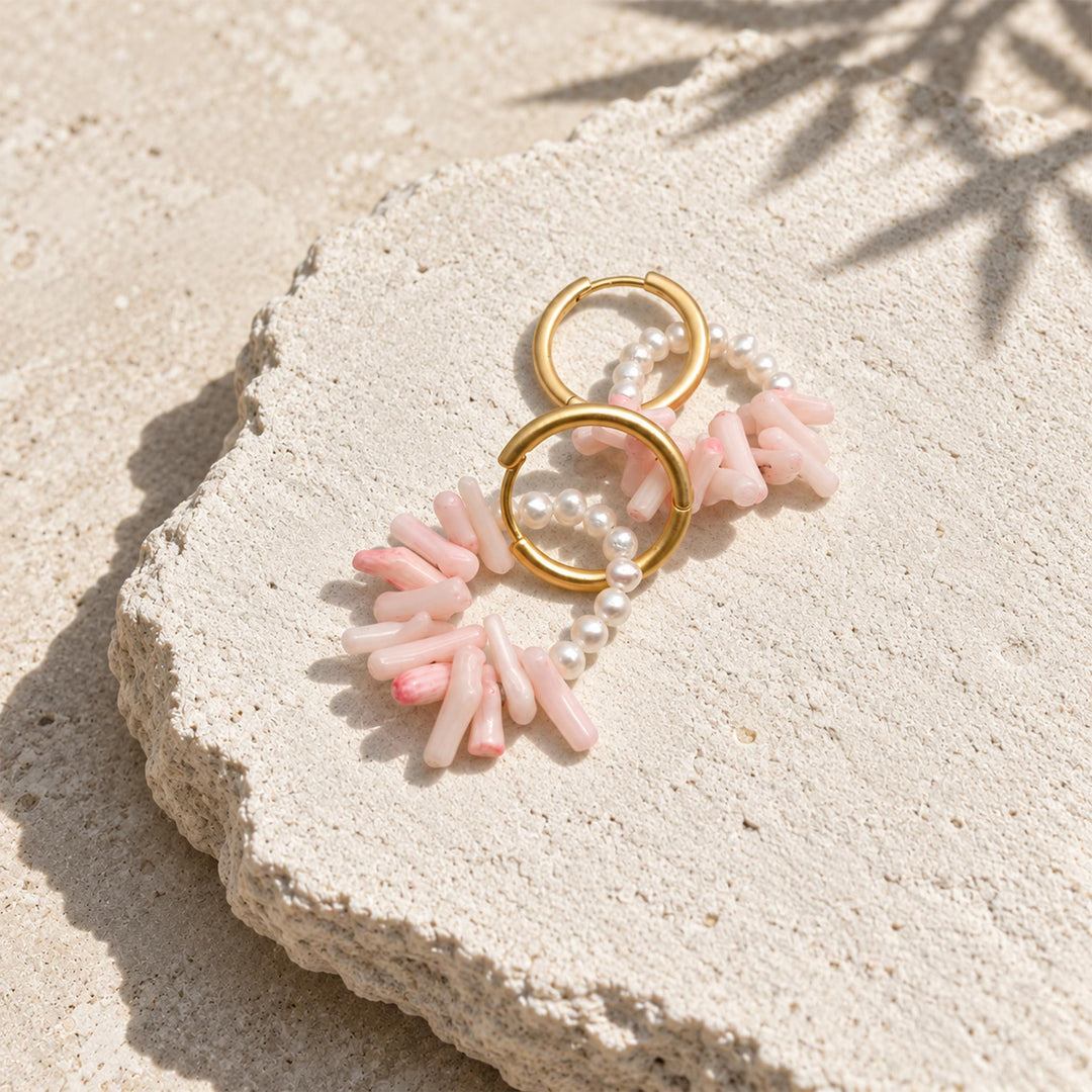 A pair of gold hoop earrings with dangling pink coral beads and freshwater pearls strands, arranged on a textured light stone surface in bright sunlight.