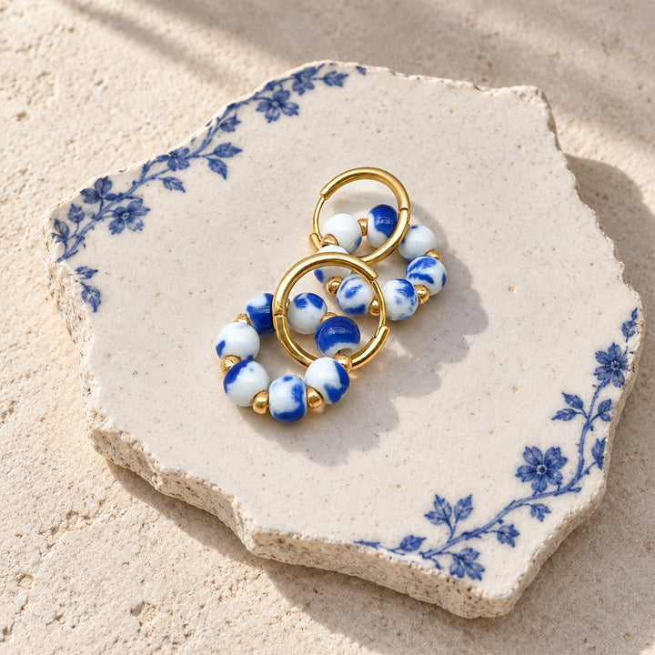 A pair of gold-tone hoop earrings with marbled blue and white ceramic beads, displayed on a decorative blue-and-white ceramic tile over a sandy beige surface.