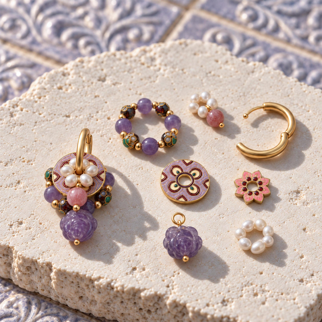 A styled flat-lay of gold-plated jewelry charms and earrings arranged on a textured beige stone slab. The jewelry features purple amethyst beads, strawberry quartz, and gold-toned metal components, including a hand-carved amethyst rose, a circular floral charm, a pearl hoop, and a gold huggie earring.