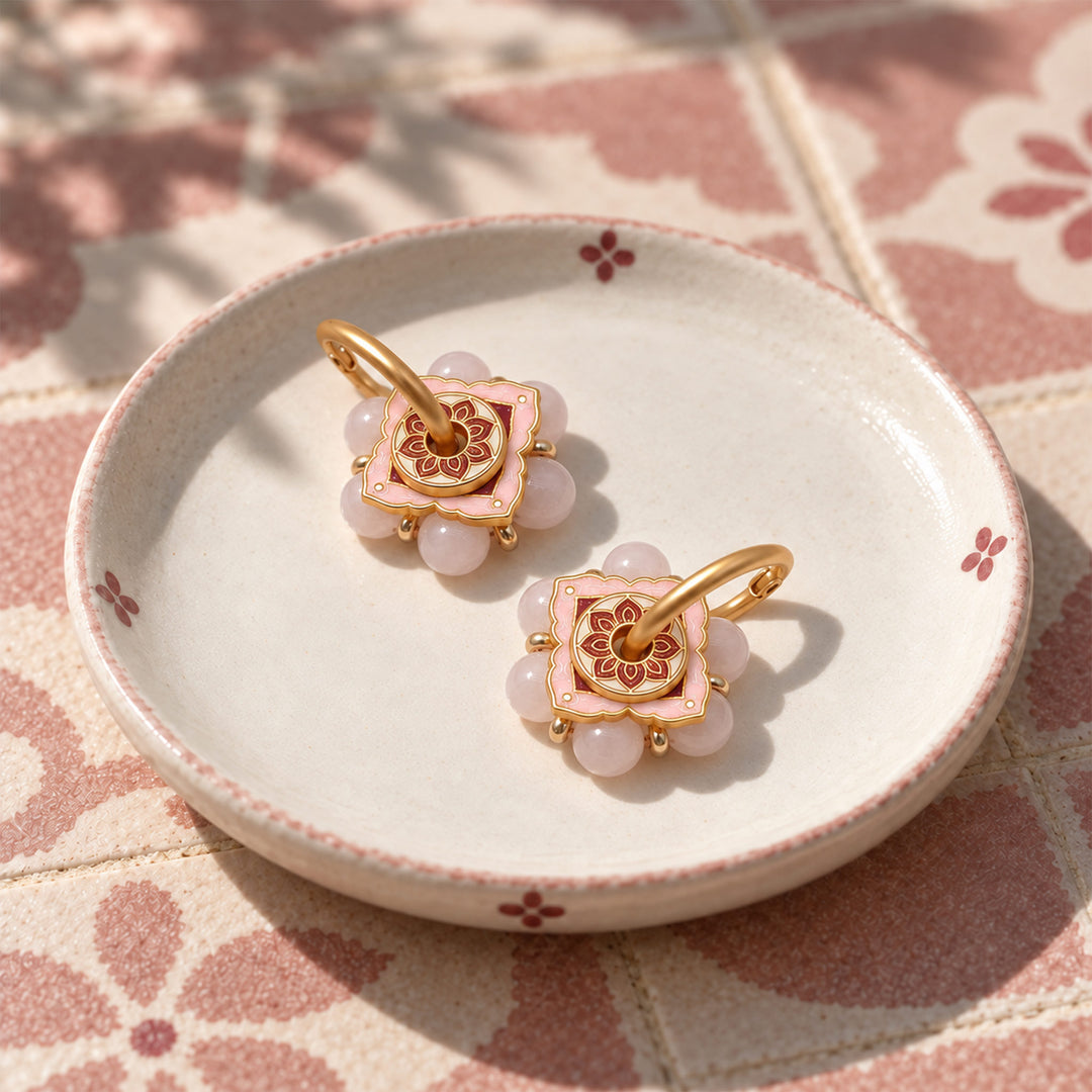 A pair of gold-toned floral hoop earrings with pink enamel and pink quartz accents, displayed on a small white ceramic dish over a pink patterned surface.