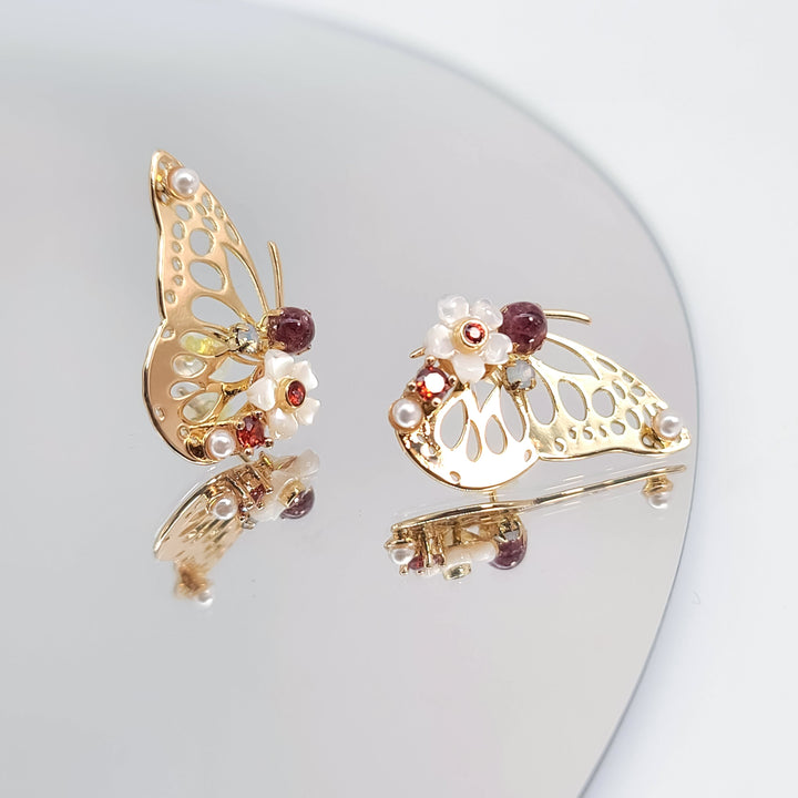 Gold butterfly earrings with red gemstones, white flowers, and pearl details on reflective surface.