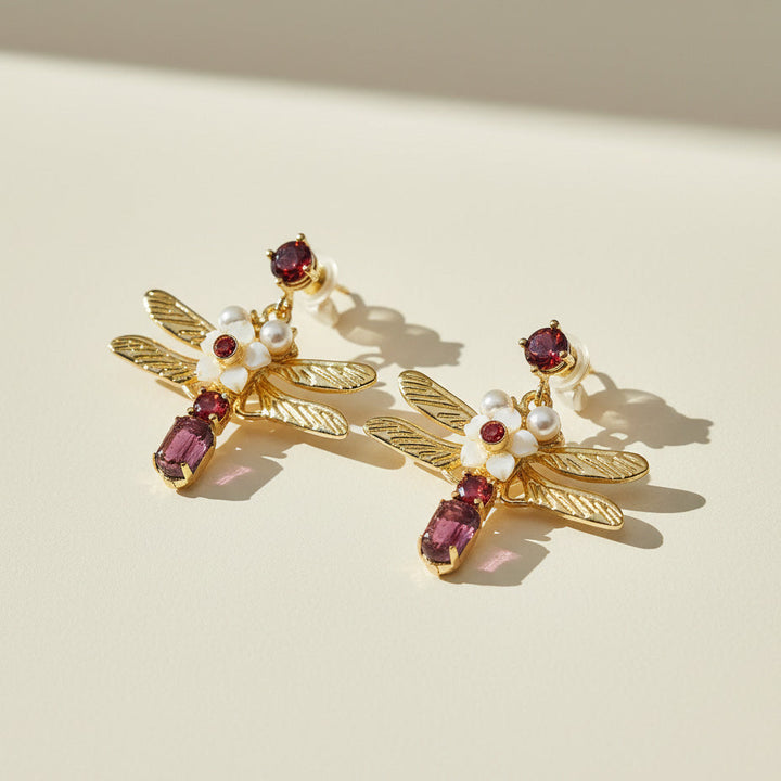 Gold dragonfly-shaped earrings with gemstones and pearls on a beige background