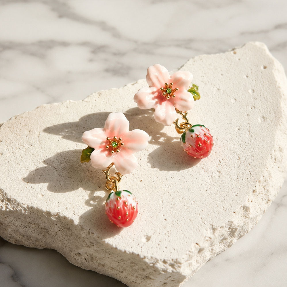 Hand-painted enamel strawberry flower drop earrings with pink cherry blossom crystals and a textured resin strawberry charm on a white stone background.
