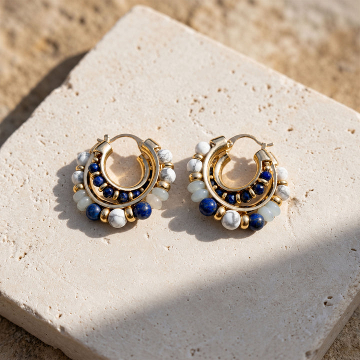 Mixed gemstone hoop earrings with blue, white and pale green beads in a gold-plated brass setting displayed on stone background
