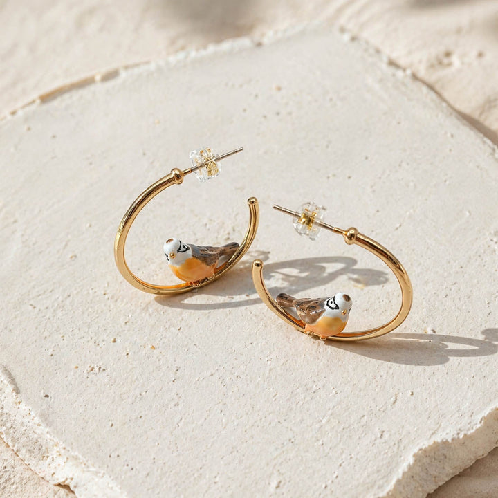 Gold hoop earrings with bird designs on a textured beige surface