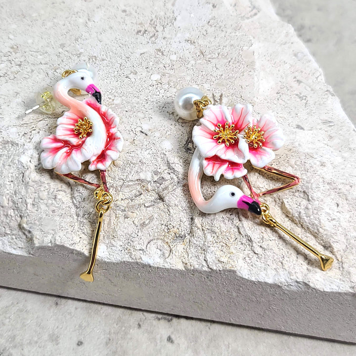 Handcrafted flamingo earrings with enamel pink flowers, pearl stud tops, and gold-tone legs, displayed on a textured beige stone background.