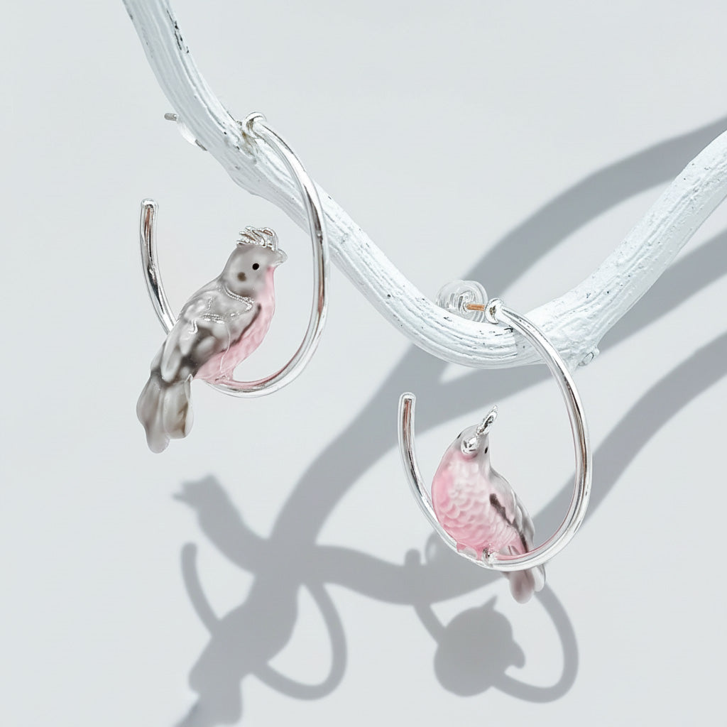 Crafted silver hoop earrings with pink and gray bird design, enamel painted cockatoo earrings on white branch display.