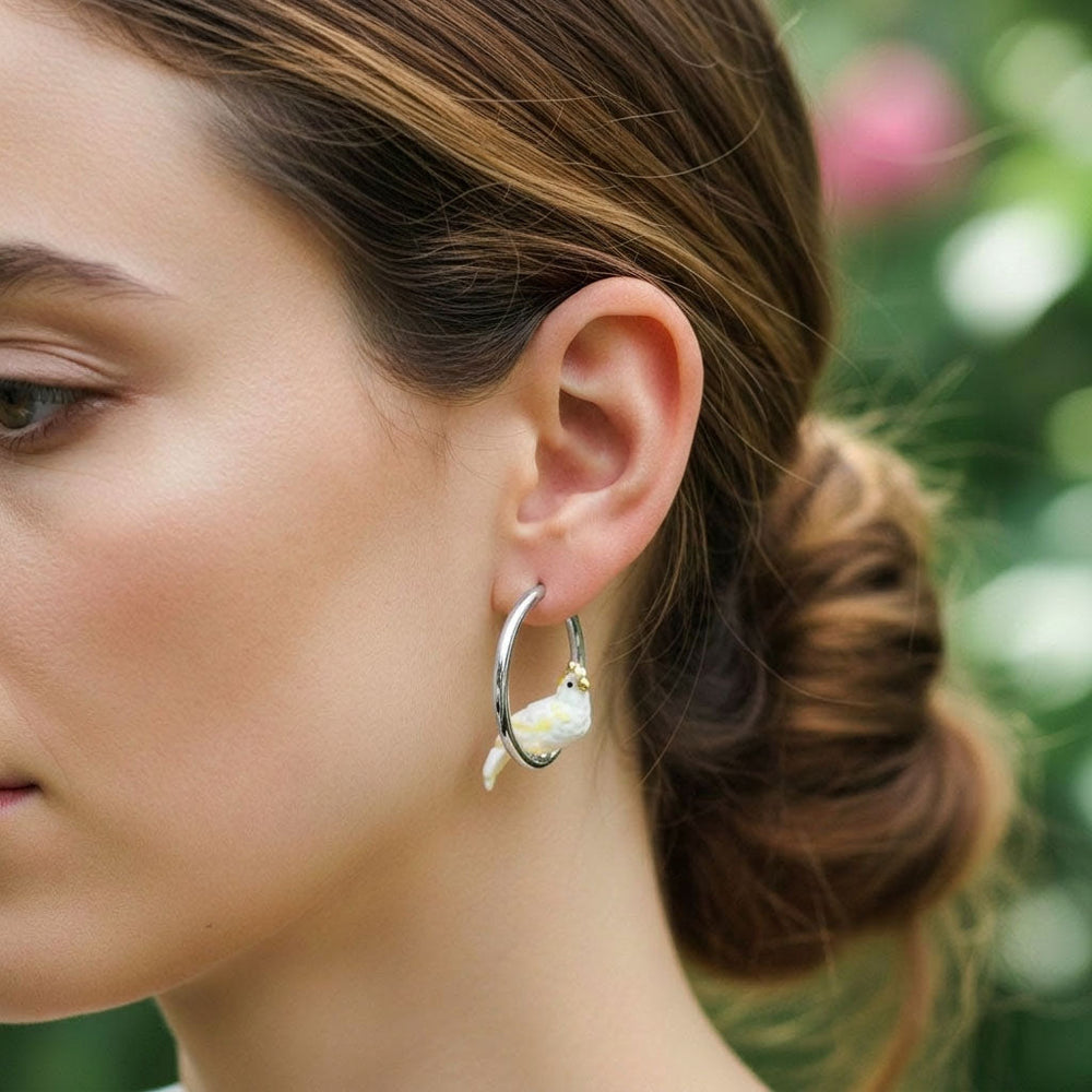 Close-up of a woman wearing a hoop earring with a small cockatoo charm against a blurred natural background.