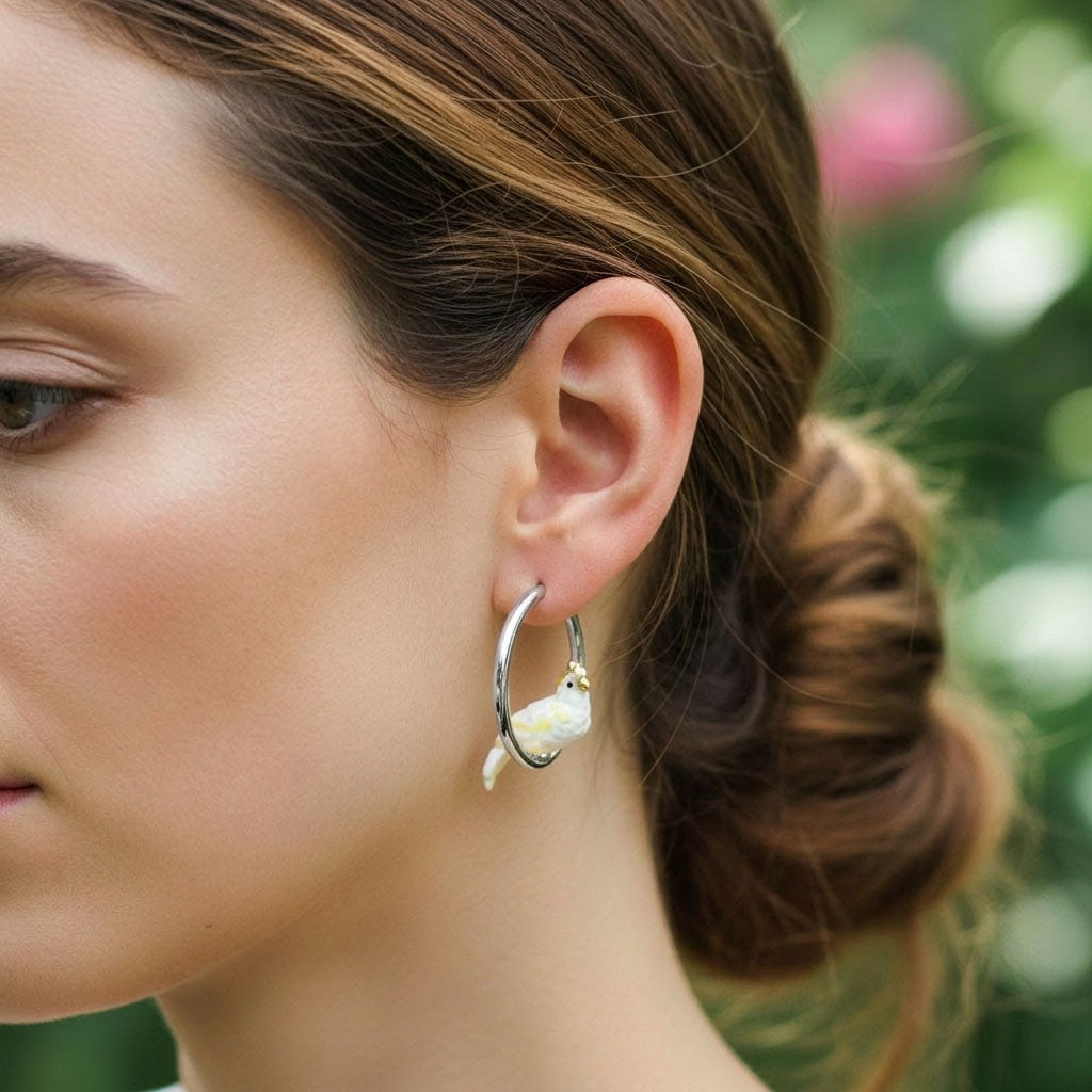 Close-up of a woman wearing a hoop earring with a small cockatoo charm against a blurred natural background.