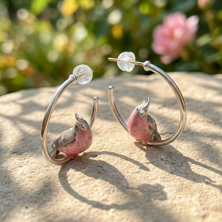 Earrings with pink and gray parrot designs on a stone surface with a blurred natural background