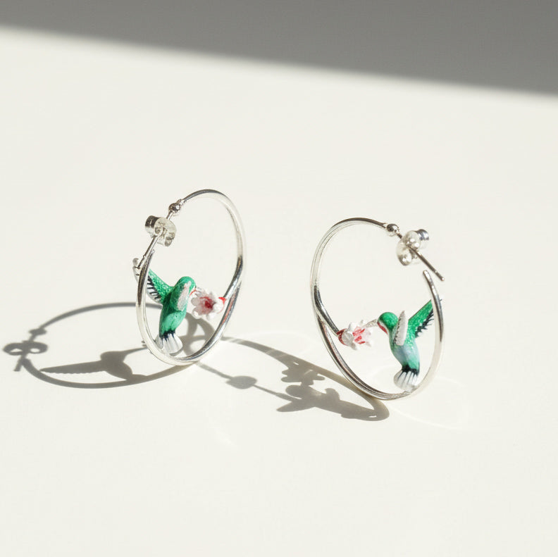 Silver hoop earrings with green enamel hummingbirds sipping from pink-white flowers, elegant crafted jewelry on light background.