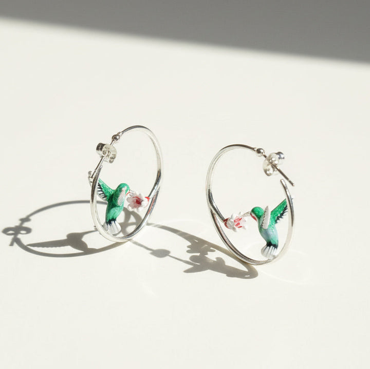 Silver hoop earrings with green enamel hummingbirds sipping from pink-white flowers, elegant crafted jewelry on light background.