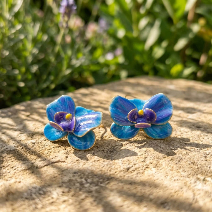 Blue orchid-shaped earrings on a stone surface with greenery in the background