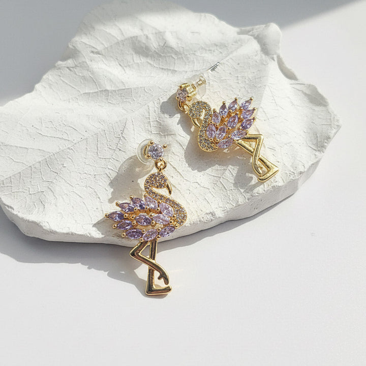 Close-up of gold flamingo drop earrings with purple CZ stones on textured white surface