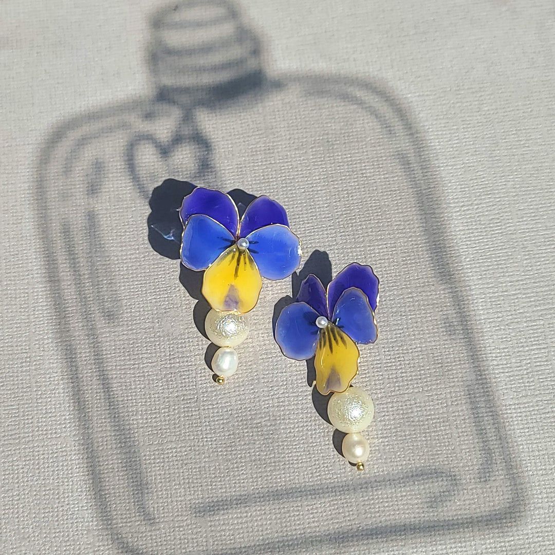 Blue and yellow pansy flower earrings with pearl drops, hand-painted enamel design captured in natural sunlight.