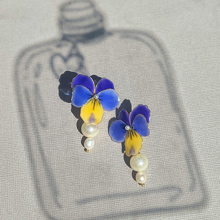 Blue and yellow pansy flower earrings with pearl drops, hand-painted enamel design captured in natural sunlight.