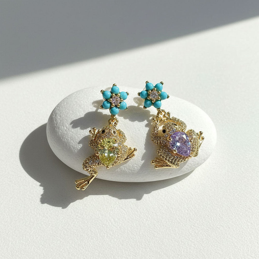 Mismatched frog and flower earrings with yellow and purple gems on white stone.