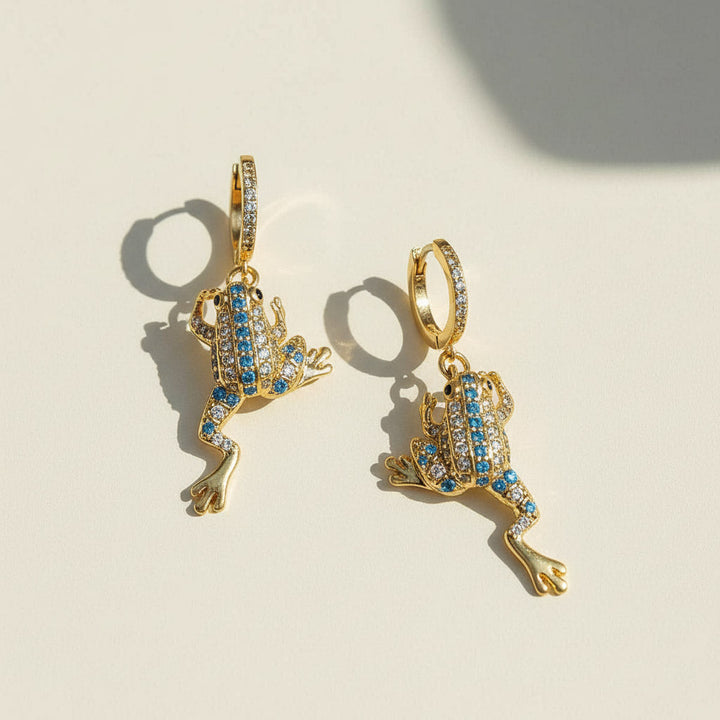 Gold frog-shaped earrings with blue gemstones on a beige background