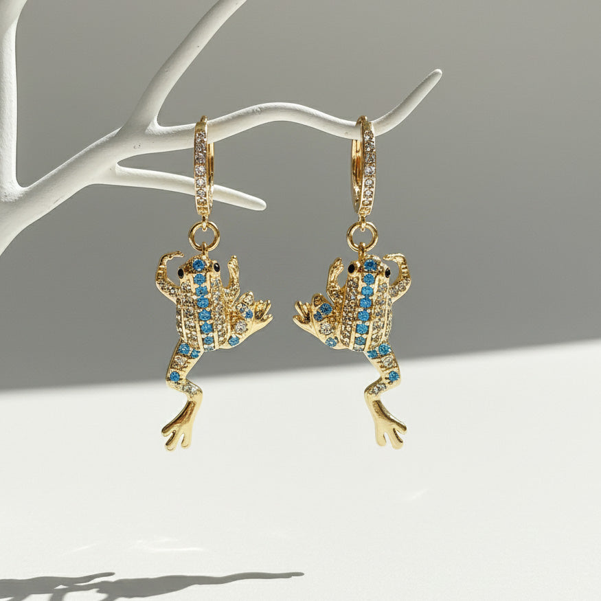 Gold frog-shaped earrings with blue gemstones on a white stand against a gray background