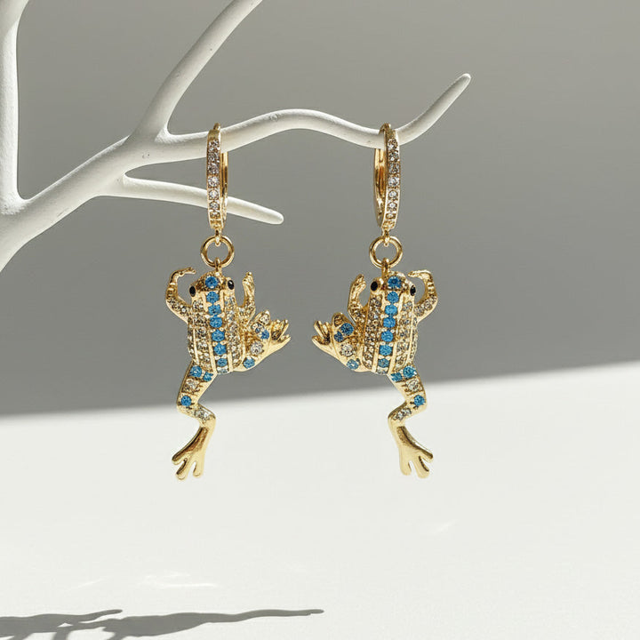 Gold frog-shaped earrings with blue gemstones on a white stand against a gray background