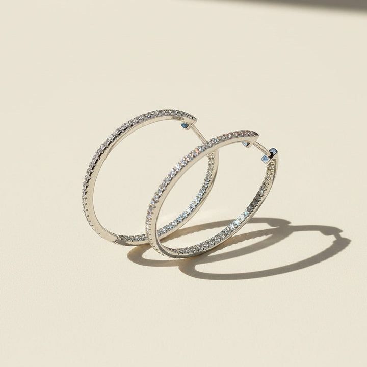 Standing display of 30mm platinum-plated moissanite hoop earrings with full pavé D/VVS1 stones on beige background.