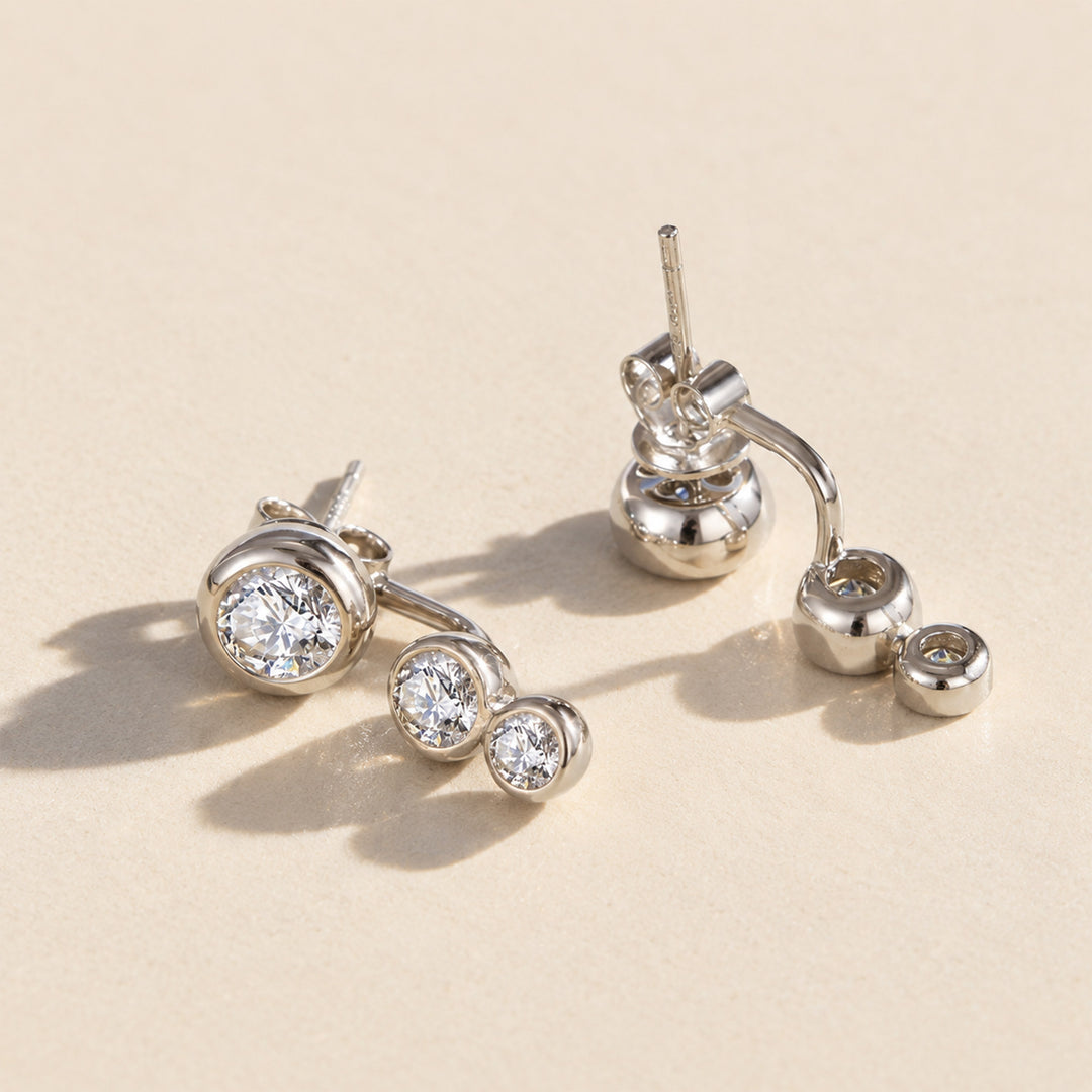 Silver three-stone moissanite ear jacket earring, shown on a warm beige background. One earring is displayed from the front showing the round bezel setting and post, while the other is shown from the side/back profile.