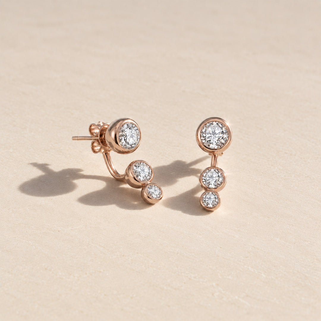 Rose gold three-stone moissanite ear jacket earrings with graduated round stones. displayed on a light beige textured surface.